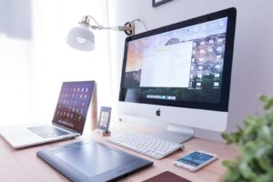 A laptop computer, drawing pad, desktop computer with keyboard, and a mobile phone all resting on a wooden desk.