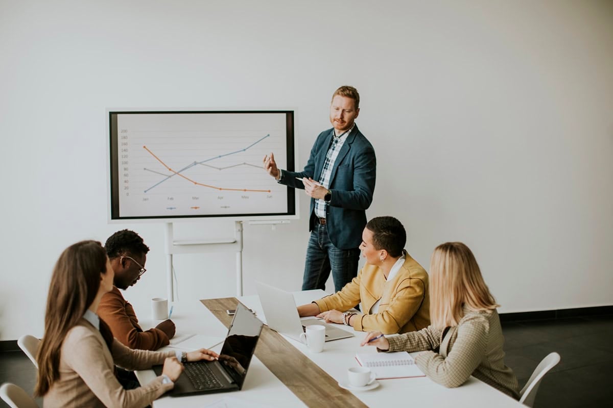A man presents a graph to four colleagues seated at a table with laptops in a meeting room.